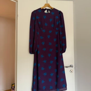 Lila blommig långklänning Fashion Union - Långklänning från Fashion Union i lila med blått blommönster. Klänningen har långa ärmar med resår vid handlederna, rund halsringning och snygg öppen rygg med knytning i nacken. Perfekt för dig som vill sticka ut med färg och mönster.