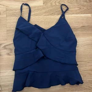 Marinblå volangtop med smala axelband - Snygg marinblå linnetopp med volangdetaljer och smala justerbara axelband. Toppen har omlottkänsla framtill och en lätt, luftig känsla som passar perfekt till jeans eller kjol. Stilren och trendig design med feminin touch.