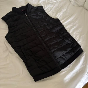 Svart quiltad dunväst från Zara Man - Svart quiltad dunväst från Zara Man med dragkedja framtill och två sidofickor. Västen har en stilren, enkel design med ribbad krage och nederkant. Perfekt att slänga över en hoodie eller tröja när det är kyligt ute.
