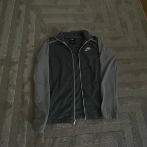Grå kofta från Nike - Snygg grå vindjacka från Nike med ljusare grå ärmar och vit Nike-logga på bröstet. Jackan har hel dragkedja framtill, ståkrage och två fickor. Perfekt för dig som gillar sportig stil och vill ha något lätt att slänga på sig.