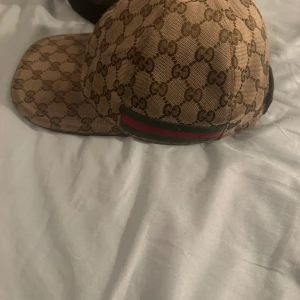 Gucci keps beige  - Snygg Gucci keps i beige och brunt med klassiskt GG-monogram över hela kepsen. Justerbar rem i brunt läder baktill och ikonisk grön-röd rand på sidan. Tillverkad i bomullsmix med exklusiv känsla, perfekt för dig som gillar streetstyle och lyx.