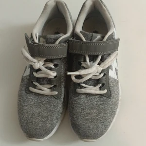 Grå sneakers med kardborre och snörning - Snygga grå sneakers med vit sula och vita detaljer. Skorna har både snörning och kardborreband för extra bra passform. Ovandelen är i mjukt textilmaterial och sidorna har en vit blixtdekal. Perfekta för en sportig och avslappnad stil.