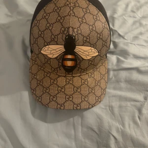 Gucci keps bee - Säljer en lyxig Gucci keps i storlek XL med klassiskt GG-monogram i beige och brunt. Framsidan har en stor bi-illustration och baksidan är i svart mesh för extra ventilation. Perfekt statement piece för dig som gillar designer och streetstyle.