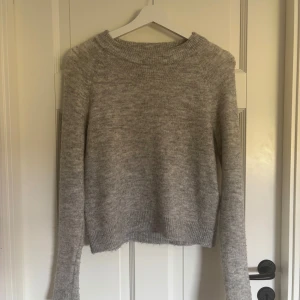 Ljusgrå stickad tröja Vero Moda - Mysig ljusgrå stickad tröja från Vero Moda i enkel och clean design. Tröjan har rund halsringning, långa ärmar och ribbade muddar. Perfekt att slänga på sig när det är kyligt ute. Passar snyggt till jeans eller kjol. På fjärde bilden kan man se tröjan i vit på hemsidan, då den har slutat säljas i grå. Den har används en del, och har ett pytte litet hål på framsidan. 