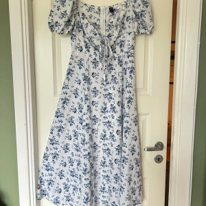 Blommig midiklänning  - Superfin midiklänning med blått blommönster på vit botten. Klänningen har puffärmar, snörning vid bysten och en romantisk v-ringning. Perfekt flowy passform och lätt material som känns luftigt och somrigt. Når mig halvvägs nedanför knäna (162 cm). Använd 1 gång, köpt second hand.