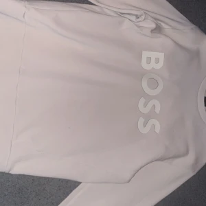 Beige BOSS tröja - Snygg beige långärmad tröja från BOSS med stor logga tryckt framtill. Tröjan har en klassisk rund halsringning och är gjord i mjuk bomull, perfekt för en clean och stilren look. Storlek M. 