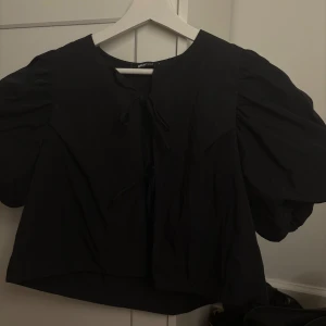Svart blus med puffärm från Gina Tricot - Svart blus från Gina Tricot med korta puffärmar och två knytningar framtill. Modellen är croppad och har en luftig passform. Perfekt för dig som gillar trendiga detaljer och enkel stil. 