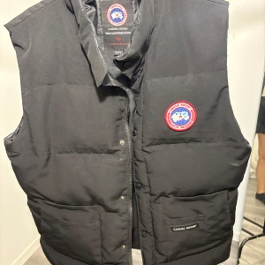 Svart dunväst från Canada Goose L/XL - Svart dunväst från Canada Goose i klassisk modell med hög krage och broderad logotyp på bröstet. Västen har dragkedja och knappar framtill samt två stora fickor. Sömmen har gått upp lite i armhålan men det går att åtgärda enkelt. 