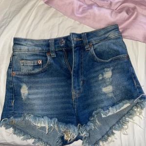 Blå jeansshorts med fransig kant - Snygga blå jeansshorts i storlek 32 med hög midja och slitna detaljer framtill. Shortsen har fransig kant nertill och klassiska fickor både fram och bak. Från divided! 