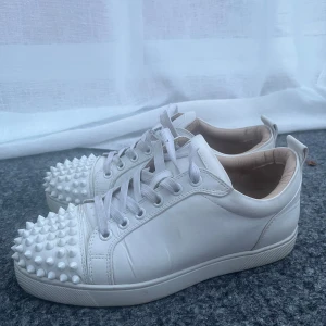Vita Louboutin sneakers med nitar - Snygga vita sneakers från Louboutin i skinn med coola vita nitar över tån och klassisk röd sula. Skorna har snörning och en clean, modern look som sticker ut. skosnörena går att ta av och tvätta om det skulle behövas 🥰 ( lådan till skorna finns och kommer skickas med ) 