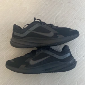 Svarta Nike sneakers storlek 40.5 - Snygga svarta Nike sneakers med mörkgrå detaljer och klassisk swoosh på sidorna. Skorna har snörning och en sportig, modern design med mesh och syntetmaterial för extra komfort. Perfekta för dig som gillar stilrena och funktionella skor.