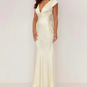 Champagnefärgad långklänning i satin - Supersnygg långklänning i champagnefärgad satin med djup v-ringning och öppen rygg med twistad detalj. Klänningen har korta ärmar och figurnära passform som ger en elegant siluett. Perfekt för dig som vill sticka ut med en lyxig look. Är i stl xl men kan passa för de i L också. Den är aldrig använd, säljs då jag inte kan ha den 