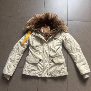 Parajumpers beige vinterjacka XXS - Snygg beige vinterjacka från Parajumper. Jackan har flera fickor, gul detalj på axeln och patch på ärmen. Perfekt för kalla dagar och har dragkedja framtill. Slim fit-modell med quiltade detaljer. Passar både XXS och XS 