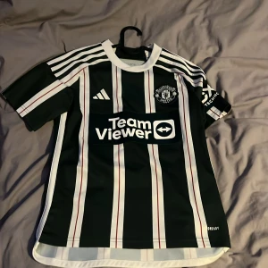 Manchester United randig fotbollströja Adidas - Snygg Manchester United fotbollströja från Adidas i svart med vita och röda ränder. Klassisk klubbmärke på bröstet, TeamViewer och DXC Technology-loggor. Kortärmad modell i lätt och ventilerande material, perfekt för match eller träning. Och nästan aldrig använd🙌
