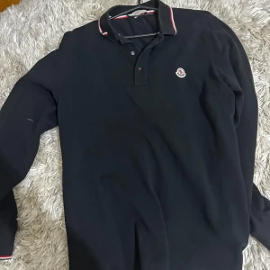 Svart långärmad pikétröja Moncler - Svart långärmad pikétröja från Moncler med klassisk krage och knäppning framtill. Kragen har snygga röda och vita ränder och Moncler-loggan sitter broderad på bröstet. Tröjan är gjord i mjuk bomull och passar perfekt till en clean och stilren look.