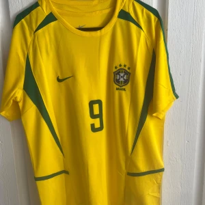 Brasilien Ronaldo 9 Nike fotbollströja - Gul Brasilien fotbollströja från Nike med gröna detaljer, nummer 9 och Ronaldo på ryggen. Klassisk design med korta ärmar, rund hals och CBF-emblem på bröstet. Perfekt för dig som älskar fotboll och vill hylla en ikonisk spelare.