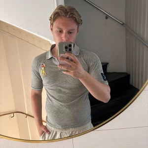 Grå pikétröja Polo Ralph Lauren M - Snygg grå pikétröja från Polo Ralph Lauren med klassisk krage och korta ärmar. Tröjan har en stor broderad logga i gult och rött på bröstet samt siffra på ärmen. Perfekt för en clean och sportig stil. Sitter mer som S