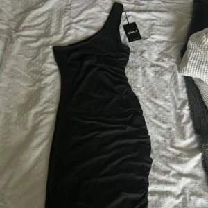 Svart omlottklänning från ONLY - Supersnygg svart klänning från ONLY som kan bäras både som one shoulder och halterneck. Klänningen är figurnära och har en figursydd passform. Går ungefär till knät. Med prislapp kvar!  Väldigt svalt och lent bomullsmaterial.