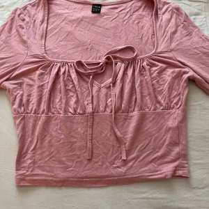 Rosa långärmad topp med knyt från SHEIN - Supersöt ljusrosa långärmad topp från SHEIN med fyrkantig ringning och knytband framtill. Toppen har en lätt rynkad detalj vid bysten och är croppad i modellen. Perfekt för dig som gillar en feminin och trendig stil.