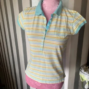 Randig polotopp i pastellfärger - Snygg randig polotopp med korta ärmar och pastellfärgade ränder i gult, blått och vitt. Kragen och knappslån är i ljusblått och toppen har en liten bröstficka. Perfekt för dig som gillar en fräsch och färgglad stil.