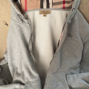 Grå zip  från Burberry - Snygg grå zipfrån Burberry med klassiskt rutigt foder i huvan. Jackan har dragkedja framtill, två fickor och är fodrad med mjuk fleece. Perfekt för dig som vill ha en stilren och bekväm jacka med exklusiv känsla.dragkedjan är trasig så den går inte att stänga.