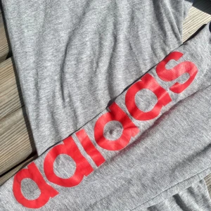 Grå leggings från Adidas  - Leggings från Adidas med stor röd logga längs vänstra ena benet och klassisk Adidas-logga i grå vid höften. Mjukt och stretchigt material, perfekt för träning eller chill. Tight passform och elastisk midja för extra komfort. Inga defekter.❣️✨