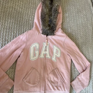 Rosa hoodiejacka GapKids med fuskpäls - Mysig rosa hoodiejacka från GapKids med stor vit GAP-logga framtill. Jackan har dragkedja, två fickor och huva fodrad med vit fuskpäls samt pälsdetalj runt huvan. Perfekt för kyliga dagar och riktigt skön att ha på sig.
