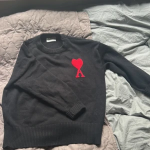 Svart stickad tröja från Ami Paris - Svart stickad tröja från Ami Paris med röd broderad hjärtlogga och bokstaven A på bröstet. Klassisk rund halsringning, ribbade muddar och nederkant. Perfekt för dig som gillar stilrena plagg med en twist.