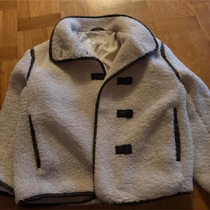 Vit teddyjacka med svarta detaljer - Säljer en vit teddyjacka med svarta kantband och stora knappar framtill. Jackan har en oversized krage, långa ärmar och två fickor med svarta detaljer. Perfekt för dig som vill ha en mysig och trendig look under kyliga dagar.