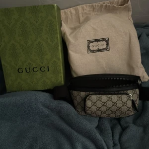 Gucci beige magväska med GG-mönster - Säljer en snygg magväska från Gucci i beige canvas med klassiskt GG-mönster och svarta skinn- och textildetaljer. Väskan har justerbart band, dragkedja och en extra ficka framtill. Perfekt för att bära det viktigaste med stil.