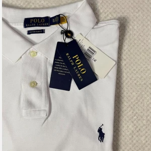 Vit pikétröja från Polo Ralph Lauren - Klassisk vit pikétröja från Polo Ralph Lauren med blå broderad logga på bröstet. Tröjan har krage, knappar framtill och är tillverkad i bomull. Perfekt för en clean och stilren look.