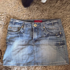 Jeanskjol från Pop Chic med nitar - Snygg ljusblå jeanskjol från Pop Chic med coola slitningar och dekorativa nitar på fickorna. Klassisk femficksmodell med knapp och dragkedja framtill. Broderade detaljer på bakfickorna. Perfekt för dig som gillar en trendig och avslappnad stil.