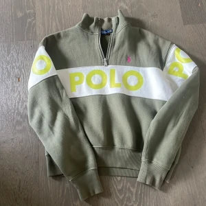 Olivgrön half zip tröja från Polo Ralph Lauren - Olivgrön långärmad tröja från Polo Ralph Lauren med vit och neongul POLO-logga över bröst, rygg och ärmar. Tröjan har half zip, ribbade muddar och en liten rosa broderad logga på bröstet. Perfekt för dig som gillar streetwear och snygga detaljer. oversized så snygg❤️