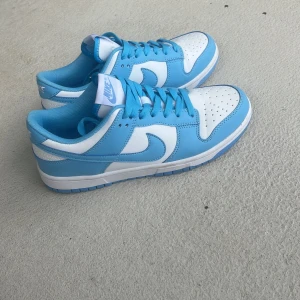 Nike Dunk Low vit/blå sneakers - Nike Dunk Low sneakers i fräsch vit och ljusblå färg. Klassisk siluett med perforerad tå, snörning och låg profil. Ovandel i skinn med blå overlays och vit bas, samt matchande blå sula och innerfoder. Använda men i mycket fint skick 
