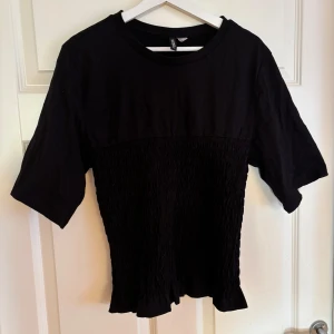 Svart topp med smock från H&M - Svart topp från H&M med smockad midja och volangkant nertill. Modellen har rund halsringning och halvkort ärm. Överdelen är slät medan nedre delen är smockad för en cool textur. 