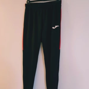 Svarta träningsbyxor från Joma M - Svarta träningsbyxor från Joma med röda sidopaneler och vit logga på låret. Byxorna har elastisk midja och är tillverkade i ett lätt och smidigt syntetmaterial som passar perfekt för sport och träning.  Helt & fint skick