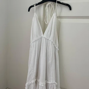 Vit långklänning  - Superfin vit långklänning från KappAhl med smock i ryggen, volangdetaljer och tunna knytband i halsen. Klänningen har v-ringning och är ärmlös, perfekt för varma dagar. Luftigt material som känns skönt mot huden.