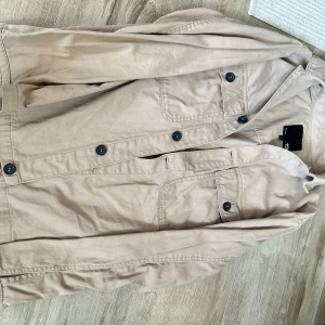 Beige overshirt från 157, storlek S - Snygg beige overshirt från 157 med klassisk krage och svarta knappar. Två bröstfickor med knappstängning ger en chill och stilren look. Perfekt att slänga över en t-shirt för en avslappnad vibe.