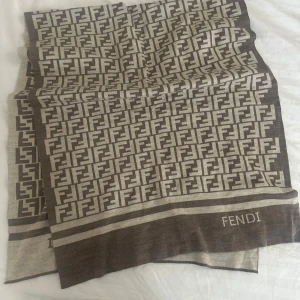 Halsduk med logomönster - Snygg halsduk i beige och brunt med logomönster över hela tyget. Rektangulär form och diskret. Perfekt accessoar för att lyfta din outfit med lyxig känsla. Tjockt tyg