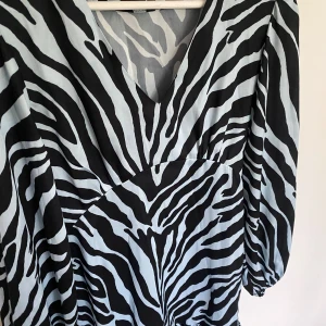 Zebramönstrad klänning med v-ringning - Snygg klänning med svartvitt zebramönster och v-ringad hals. Modellen har långa ärmar med lätt puff och resår vid ärmslut. Materialet känns mjukt och följsamt, perfekt för dig som vill sticka ut med en cool look.