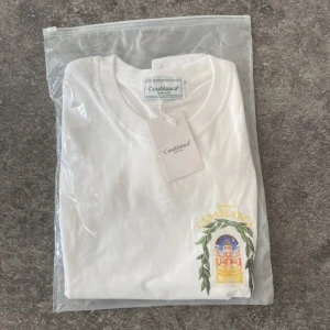 Vit t-shirt från Casablanca M - Vit t-shirt från Casablanca Tennis Club med färgglatt tryck av byggnad och blad på bröstet och ryggen. Klassisk rund hals och korta ärmar. Skön bomullskvalitet och snygg streetstil med unik grafik. Perfekt för dig som gillar trendiga prints och clean look.