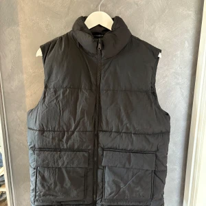 Svart dunväst med stora fickor - Snygg svart dunväst med hög krage och två stora fickor framtill. Västen har dragkedja hela vägen upp och en quiltad design som ger en cool och sportig look. Perfekt att slänga över en hoodie eller tröja när det är kyligt ute.