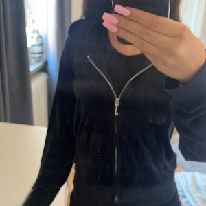 Svart velourjacka Juicy Couture - Svart velourjacka från Juicy Couture med dragkedja och huva. Klassisk broderad JC-logga på bröstet och ikonisk J-formad dragkedja. Mjuk och skön, perfekt för en chill och trendig look.