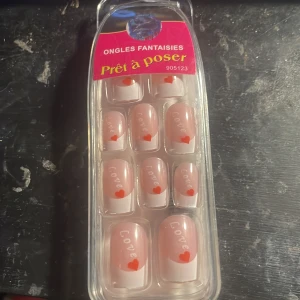 Fejk naglar med hjärta och text - Säljer ett set med lösnaglar i ljusrosa med vit tipp, dekorerade med röd hjärtdetalj och texten 'Love' på varje nagel. Naglarna är i olika storlekar och kommer i originalförpackning. Perfekt för en söt och romantisk look.