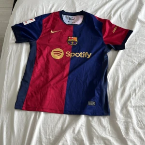 FC Barcelona Lamine Yamal matchtröja M - FC Barcelona matchtröja med Lamine Yamal 19-tryck, officiella La Liga-märket på ärmen och sponsorer som Spotify och UNHCR. Tröjan är kortärmad, i polyester, med klassiska blå och röda fält och guldiga detaljer. Perfekt för dig som älskar Barca och vill sticka ut på läktaren.