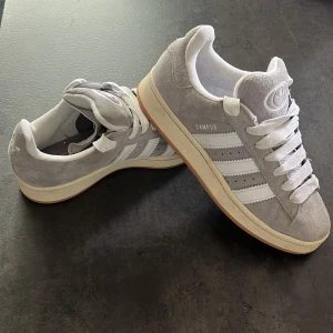Grå Adidas Campus sneakers i mocka - Snygga grå Adidas Campus sneakers i mjuk mocka med klassiska vita ränder och vit sula. Skorna har rund tå, platt sula och snörning. Perfekta för dig som gillar en clean och tidlös stil.
