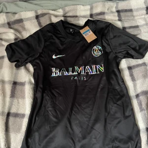 Svart PSG x Balmain fotbollströja Nike - Snygg svart PSG x Balmain Paris fotbollströja från Nike med Dri-FIT-material. Tröjan har korta ärmar, rund hals och holografiskt Balmain-tryck på bröstet. Perfekt för dig som vill sticka ut på planen eller gatan.