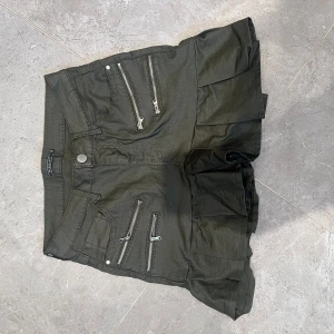 Svart kortkjol med dragkedjor - Cool svart kortkjol från The Collection med flera silvriga dragkedjor framtill och volang nertill. Kjolen har jeansinspirerade fickor både fram och bak, vilket ger en edgy vibe. Perfekt för dig som gillar detaljer och streetstyle.