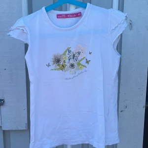 Vit topp med fjärilar från Chili - Söt vit topp från Chili med volangärmar och tryck av fjärilar och blommor framtill. Toppen har rund halsringning och är tillverkad i mjuk bomull, perfekt för en somrig look. Fina detaljer med små volanger på ärmarna ger en extra gullig touch.