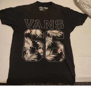 Svart Vans t-shirt med tryck - Svart t-shirt från Vans med stort tryck framtill: 'VANS 66' i vitt och beige med blommigt och palmbladsmönster. Klassisk passform och rund hals. Perfekt för dig som gillar streetwear och vill sticka ut med en snygg grafisk detalj.